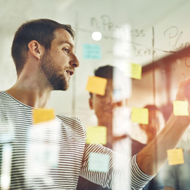 A team of product designers adjusting project plans on a glass wall with sticky notes, demonstrating collaboration, learning agility, and adaptability skills in the workplace.